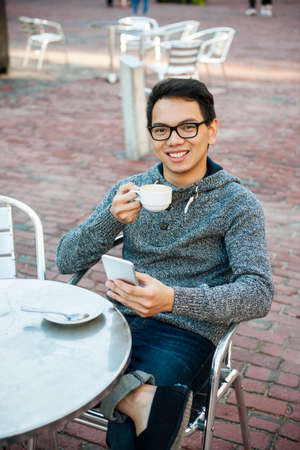 Young asian man sitting and smiling in relaxing outdoor cafe holding cup of coffee and messaging on mobile phoneの写真素材