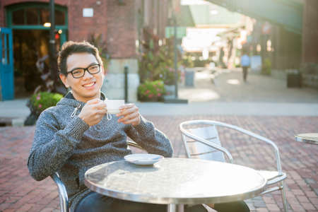 Young asian man sitting and smiling in relaxing outdoor cafe holding cup of coffeeの写真素材