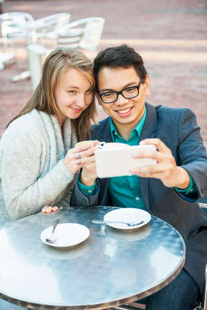 Two smiling young people looking into smartphone and taking a selfie while sitting at outdoor cafe tableの写真素材