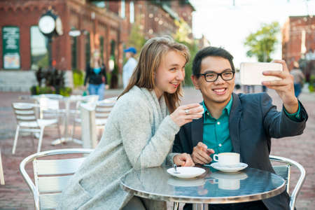 Two smiling young people taking a selfie with mobile device while sitting at outdoor cafe table on city streetの写真素材