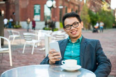 Young asian man in business casual attire sitting and smiling in relaxing outdoor cafe with cup of coffee and mobile phoneの写真素材