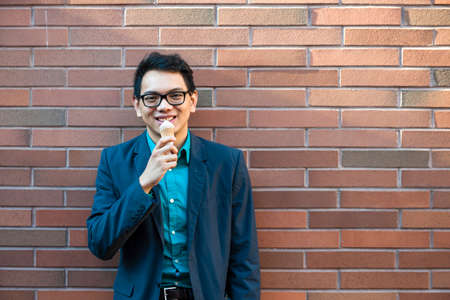 Smiling young asian man in his twenties enjoying ice cream while standing near brick wall outside on a breakの写真素材