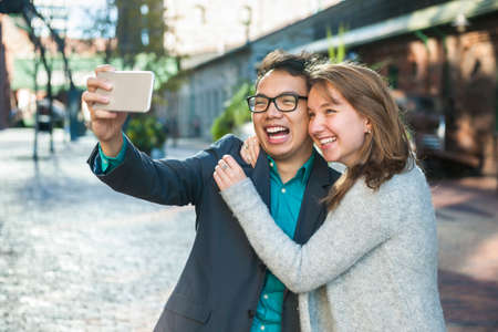 Two laughing young people taking a selfie with mobile device outside on city streetの写真素材
