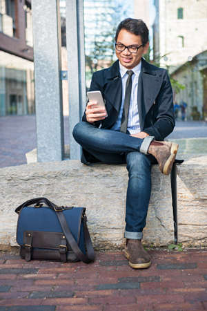 Young smiling asian business man looking at mobile phone sitting outside on city streetの写真素材
