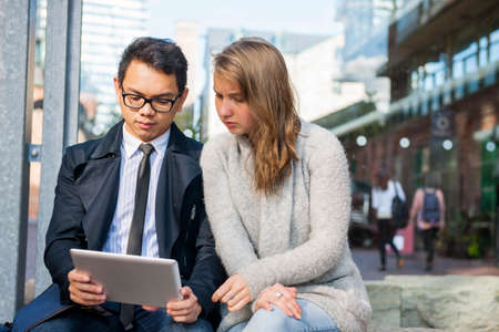 Two serious young people looking into digital tablet sitting outside on city streetの写真素材