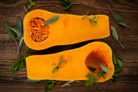 Fresh butternut squash cut in half with sage and parsley on brown wooden background, from aboveの写真素材