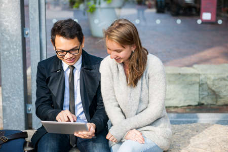 Two happy young people looking into digital tablet sitting outside on city street with copy spaceの写真素材