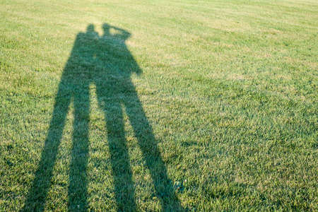 Long shadows of two people taking photos standing on grass lawn with copy space