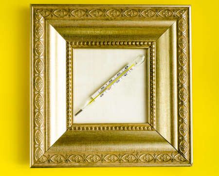 medical mercury thermometer in a gold carved frame on a yellow background.