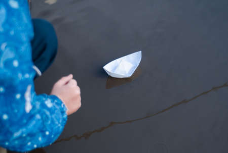 boy launches white paper boat into water. Puddles on street.