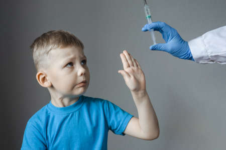 Four-year-old boy looks fearfully at the syringe in hand in doctors blue glove, in protest, he put out his palm, closing himself from the syringe.の写真素材