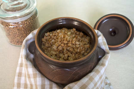 Buckwheat in a glass jar and buckwheat porridge in a clay pot wrapped with a towel on a rustic tablecloth.の写真素材