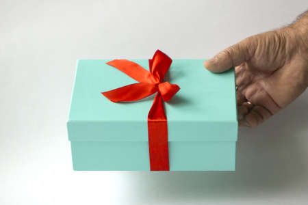 The hand of an elderly man holds out a gift in a light green box tied with a red ribbon on a white...
