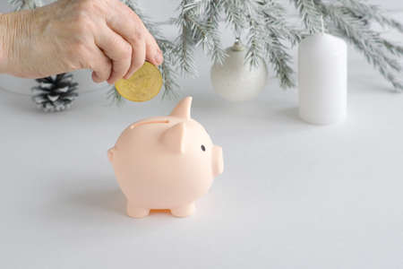 The hand of an elderly Caucasian woman lowers a metal bitcoin coin into a piggy bank pink pig on the...