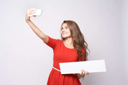 Girl makes selfie. Beautiful brunette. Red dress. White background.の写真素材