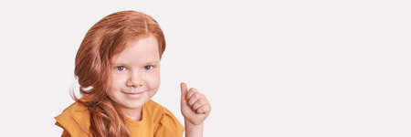 Red hair little girl. Thumb up gesture. Approve hand symbol. Female face. Looking at camera. Positive answer. Caucasian person showing agree finger. Horizontal banner. Copyspaceの写真素材