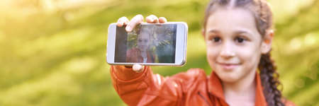 Little girl doing selfie. Happy pretty kid. Green outdoor background. Orange dress. Female person self photo. Schoolgirl phone. Travel activity. Stacation teenage influencer. Horizontal bannerの写真素材