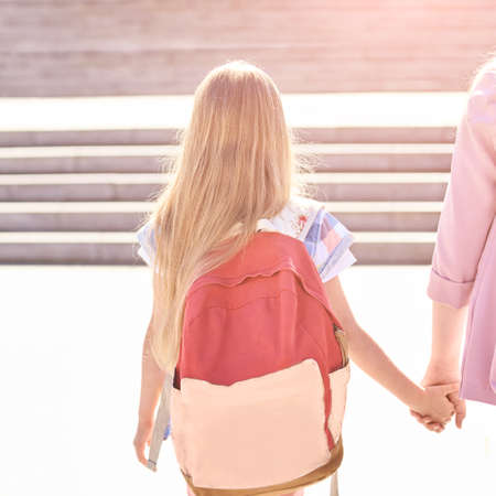 Little girl hold mother hand. Back to school. Schoolbag uniform at city. Young student. Summer education. Childhood concept. Learning in preschool. Sun light road. Squareの写真素材