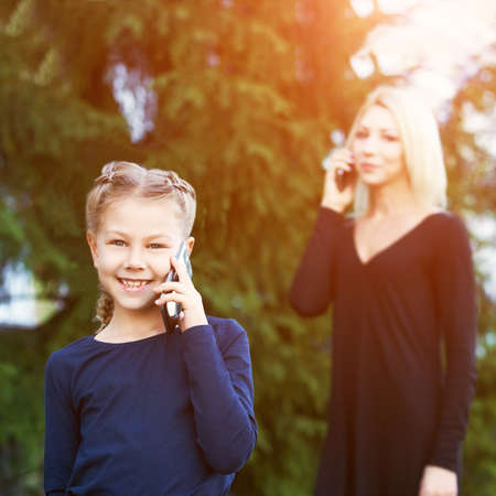 Mother and little daughter. Phone conversation. Happy family portrait. Outdoor lifestyle. Cute face. Smile positive parent. Female child love. Green background. Baby girl. Casual clothの写真素材