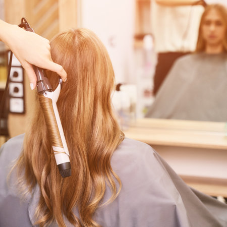 Long hair procedure. Hairstyle at salon. Woman master. Hand with iron. Adult female person. Closeup view. Studio equipment. Lady preparation. Blonde model. Mirror. Copyspace.の写真素材