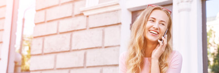 Pretty young woman talking by phone. Happy female person outdoors. Summer business portrait. Lady calling friends or boyfriend. Blonde long hair. Cute adult student at townの写真素材