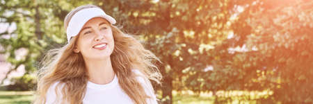 Pretty young woman wearing tennis hat. White t-shirt. Female sport player. Female person posing outdoors. Happy smiling portrait. Nature green background. Long blond hair. Wind summer dayの写真素材