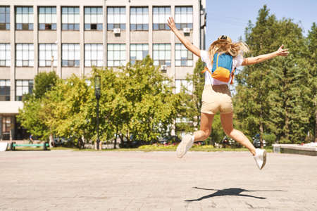 Happy young woman celebrate end of lockdown. Borders are open. Jumping at street if town. Summer time. Staycation concept. Female tourist. Backpack, hat, t-shirt, shorts. Sunny day. Back viewの写真素材
