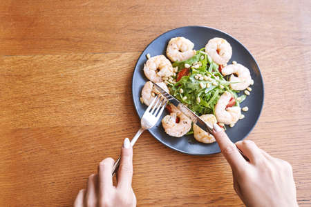 Young woman at restaurant eating shrimp salad. Health food concept. Fork and knife. Ountdoor cafe. Hands near plate. Mediterranean diet mealの写真素材