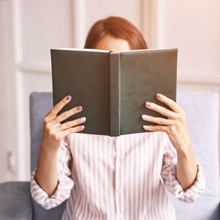 Young woman sitting at chair. Hide face behind notebook. Study concept. Safety lockdown distance teaching. Female person. Quarantine office. Business writerの写真素材