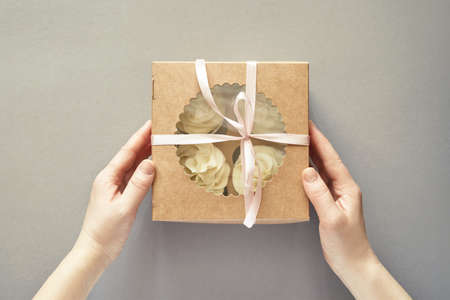 Paper box with cream cupcake. Birthday sweet bakery. Cookies four pack. Yummy buttercream dessert. Wedding party food. Grey background. Holiday recipe. Hand holdingの写真素材