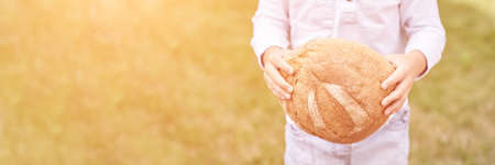 Child holds and bite round bread. Healthy food. Carrying big fresh baker bun. Rustic product. Enjoy warm breakfast. Craft cuisine. Home made lunch. Green outdoor background. Anonymous trendの写真素材
