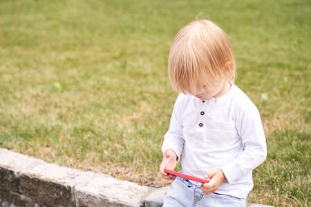 Little boy playing with smartphone. Outdoor faceless portrait. Green background. Toddler hold and...