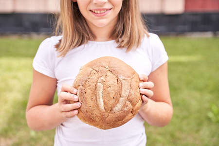 Child holds and bite round bread. Healthy food. Carrying big fresh baker bun. Rustic product. Enjoy warm breakfast. Craft cuisine. Home made lunch. Green outdoor background. Anonymous trendの写真素材
