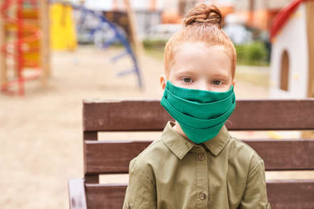 Child in safety mask. New normal.   virus safety portrait. Red hair girl. Daughter. Playground background. Outdoors. Health careの写真素材