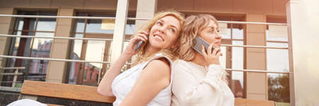 Two pretty young woman talking by phone. Happy female person outdoors. Summer city business...