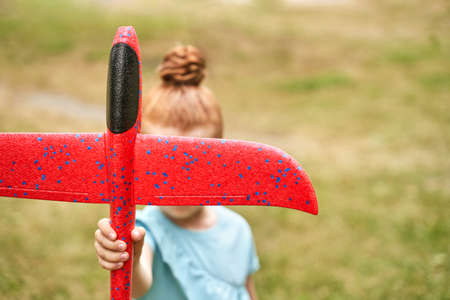 Little girl with airplane toy. Adventure fly concept. Stay home game. Family garden activity. Red hair female portrait. Styrofoam childhood pilot. Summer time. Park outdoor aviationの写真素材