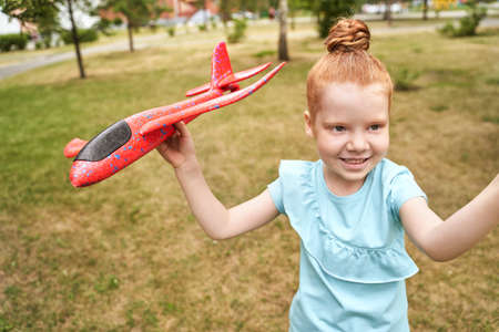 Little girl with airplane toy. Adventure fly concept. Stay home game. Family garden activity. Red hair female portrait. Styrofoam childhood pilot. Summer time. Park outdoor aviationの写真素材