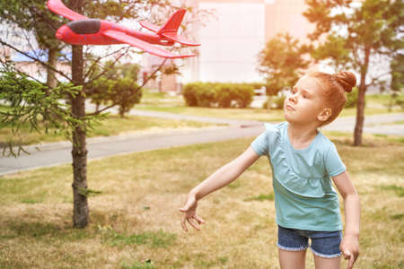 Little girl with airplane toy. Adventure fly concept. Stay home game. Family garden activity. Red hair female portrait. Styrofoam childhood pilot. Summer time. Park outdoor aviationの写真素材