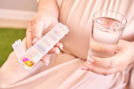 Pregnant woman eat pregnancy pills. Flu supplements. Taking allergy drugs. Medical fish omega 3 food. People body. Drink water. Mother hand. Sick female person at home. Daily tablet containerの写真素材