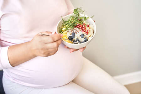 Pregnant woman eating healthy food. Organic calcium salad with fruits and microgreen. Round plate. Home dinner or lunch. Vitamin cereal. Holding bowl in hands.の写真素材