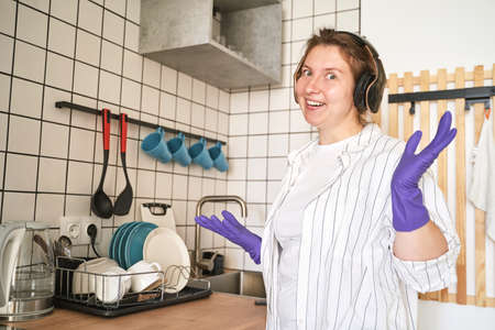 Young happy woman wash dishes at kitchen. Bright female portrait. Wow emotion. Listen music. Hand in purple gloves. Sink and platesの写真素材