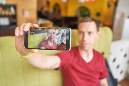 Pretty male person doing selfie photo on smartphone. Wanderlust blogger lifestyle. Happy man. Alone model. Staycation cafe concept. Face portraitの写真素材
