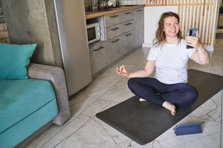 Young woman doing self portrait on yoga mat. Home vacation indoors portrait. Blogger selfie stick. Alone in interior. Happy emotion. Smiling female person. Lifestyle action. Posing influencerの写真素材