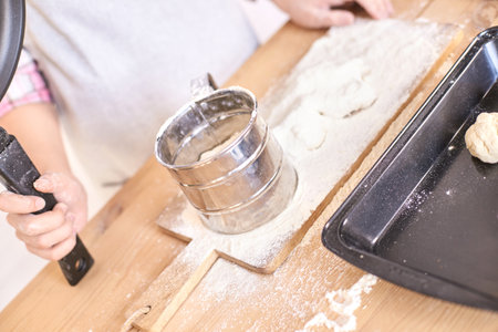 Baking preparation: child using sifter with dough on wooden board and tray in kitchen setting....