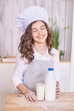 Joyful young baker in chef hat with milk bottle and glass in kitchen setting. Pretty child portrait. Attractive brunette student. Children emotion. People bakingの写真素材