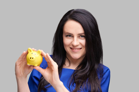 Woman smiling at camera holding yellow piggy bank in blue attire on gray background for financial conceptsの写真素材