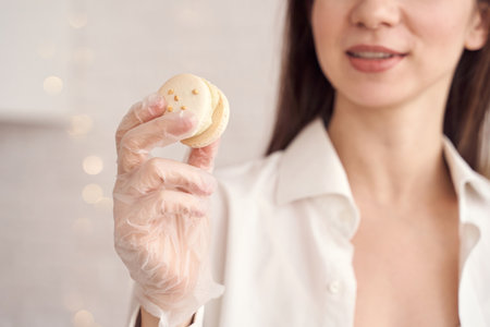 Smiling woman in white shirt holding delicious macaron with gloved hand in soft light kitchen setting.の写真素材