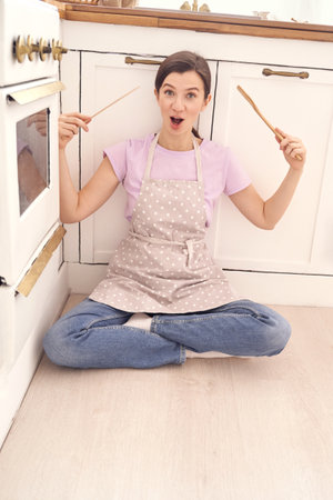 Cheerful woman in apron sitting on kitchen floor holding cooking utensils, embracing playful cooking mood.の写真素材