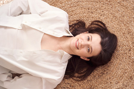 Relaxed woman lying on woven rug in cozy white shirt and casual top with a warm smile and natural light ambiance.の写真素材