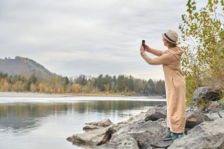 Woman capturing scenic lake view with smartphone on rocky shore in autumn wilderness.の写真素材